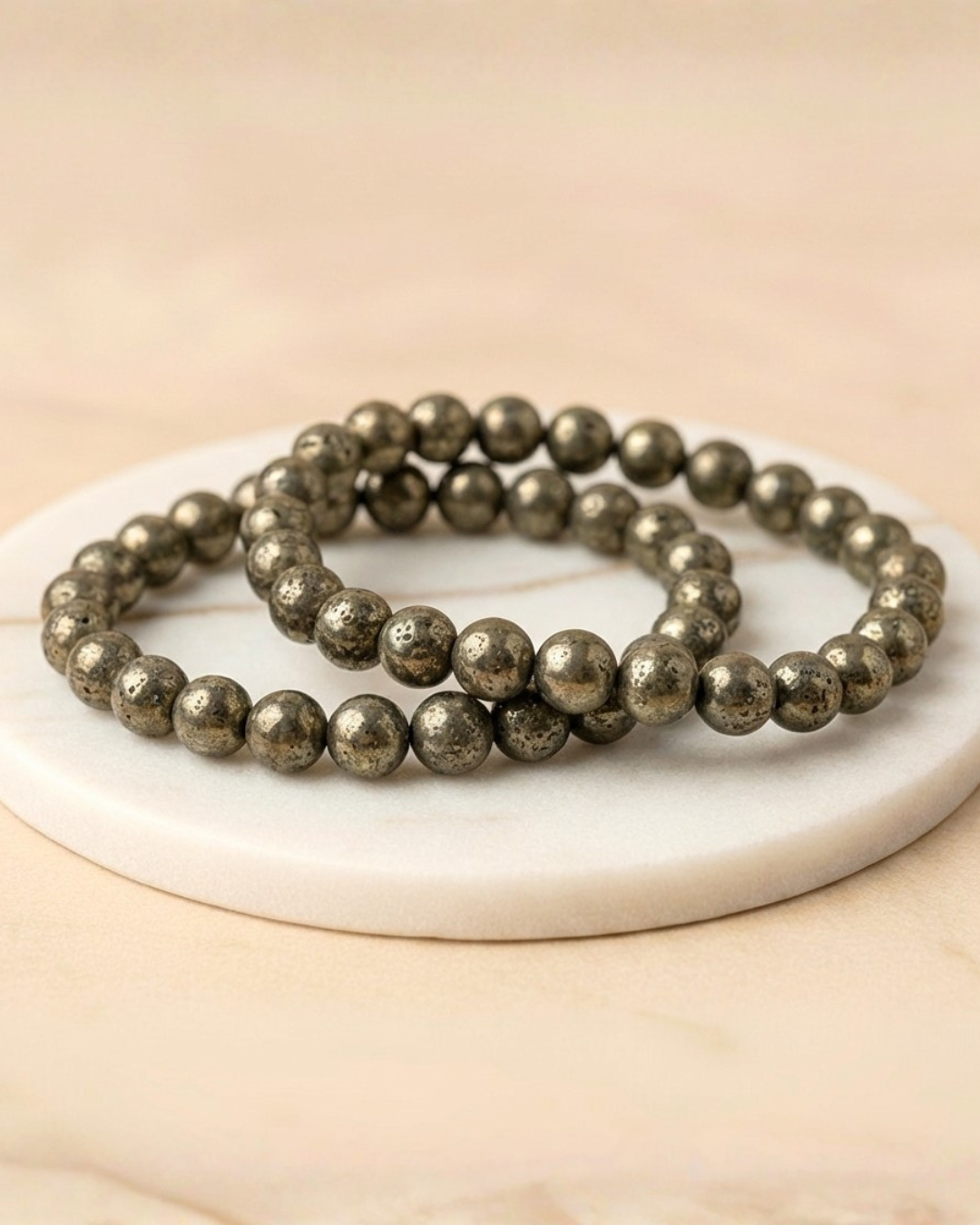 Two pyrite beaded bracelets on a white circular stone surface with a beige background