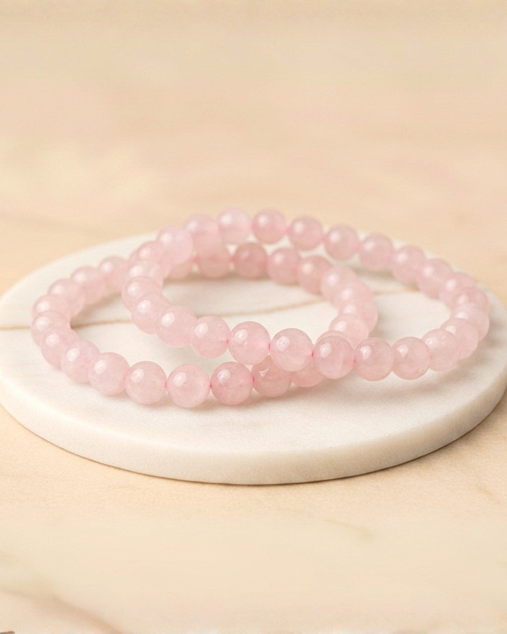 Two pink beaded Rose Quartz bracelets on a white circular base with a beige background